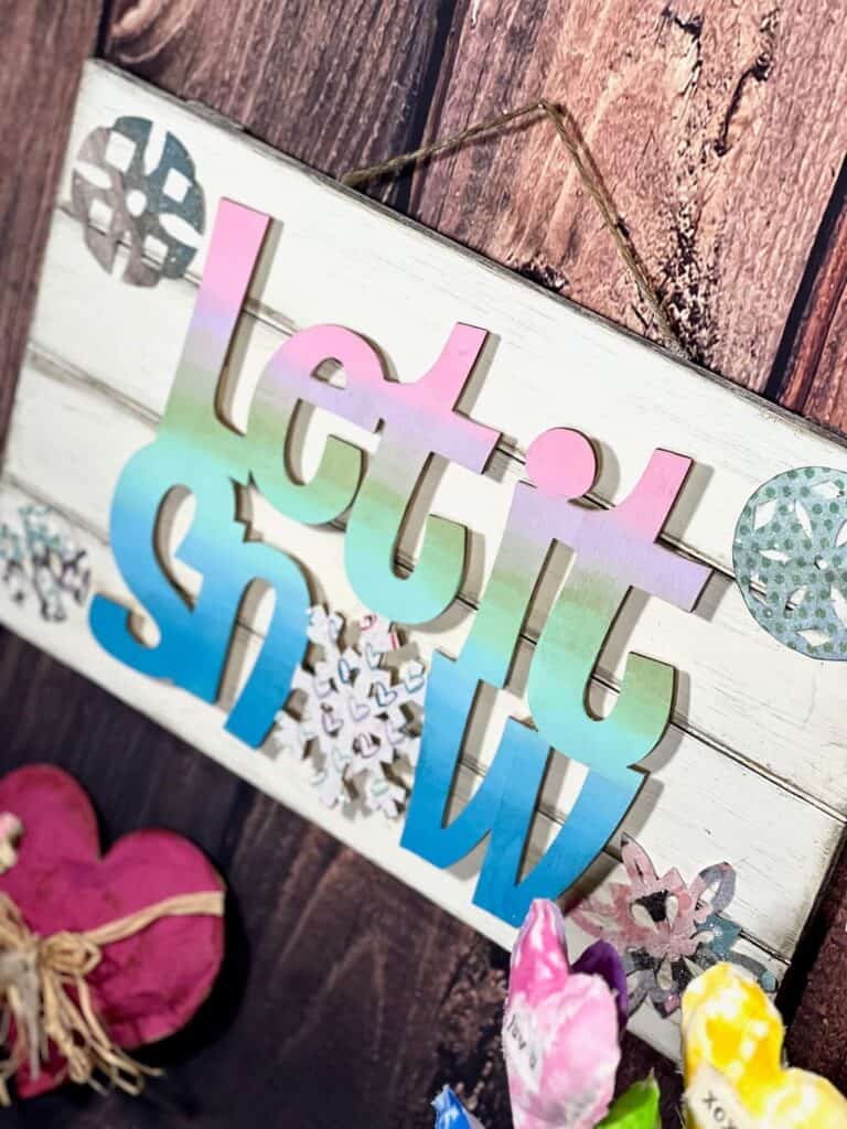Dollar Tree Let it Snow Winter Decor with the words being a pastel ombre, a white wood pallet background and colorful sparkly snowflakes. The "O" in snow is a snowflake.