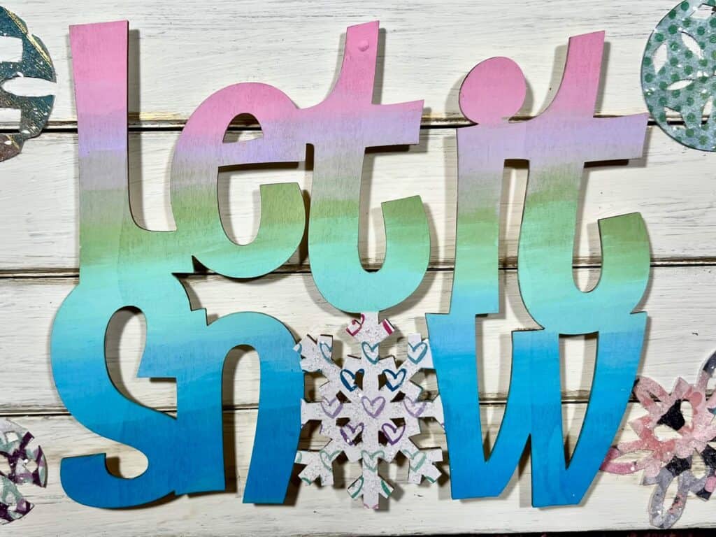 The "let it snow" painted pastel ombre from pink to purple to green to teal to blue with a snowflake for the "O" that is covered in Diamond Dust.