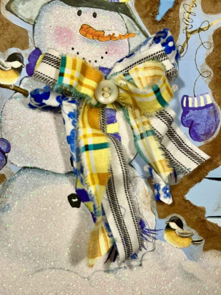 A bow made with yellow, blue and white fabric glued on top of the snowman  scarf.