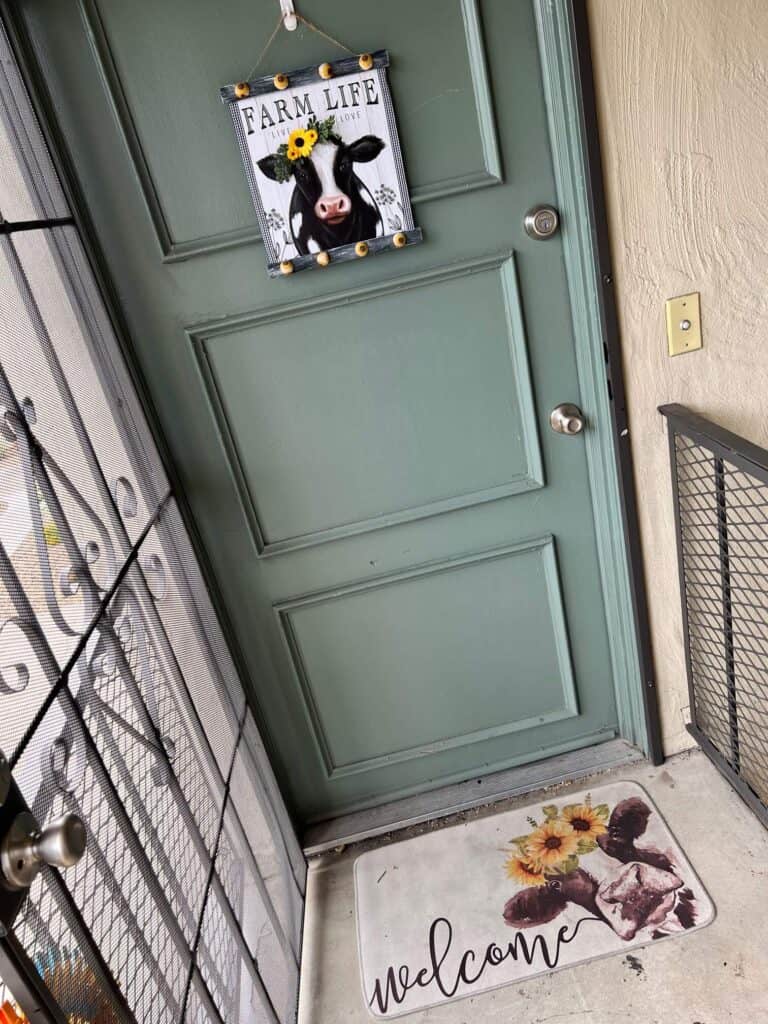 The completed Farm Life Cow Calendar frame hanging on a moss green front door with a Cow and sunflower "welcome" mat.