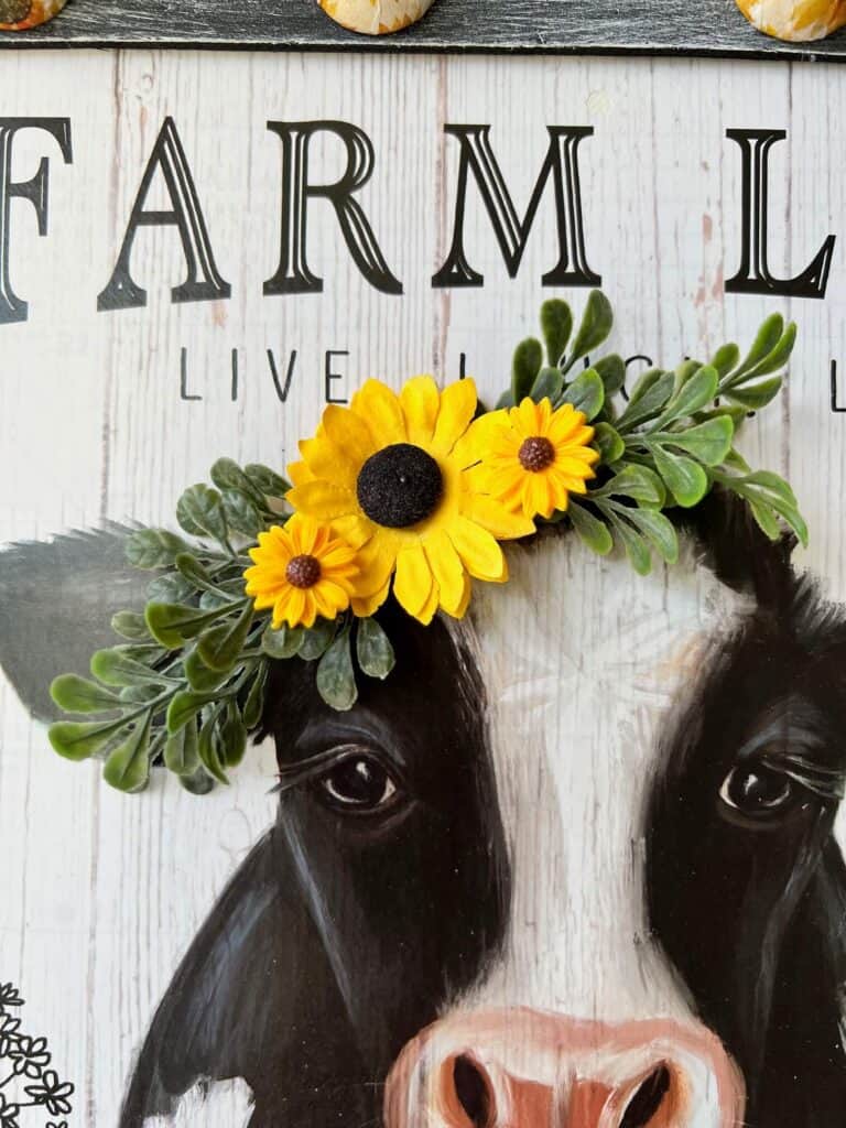 The cow calendar print has sunflowers and green boxwood glued to the top of the cows head next to its ear.
