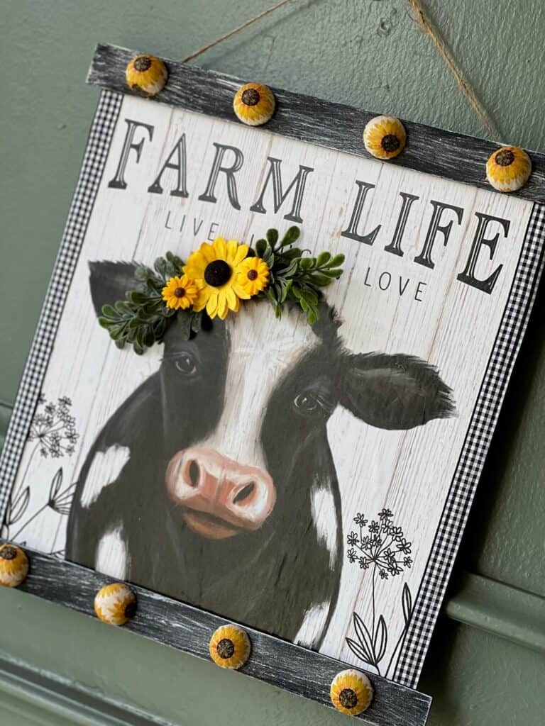 A DIY Decor frame with the Dollar Tree Farm Life Cow Print Calendar with sunflowers in the cows head, and sunflower beads on the top and the bottom of the frame, hangin with a twine hanger.