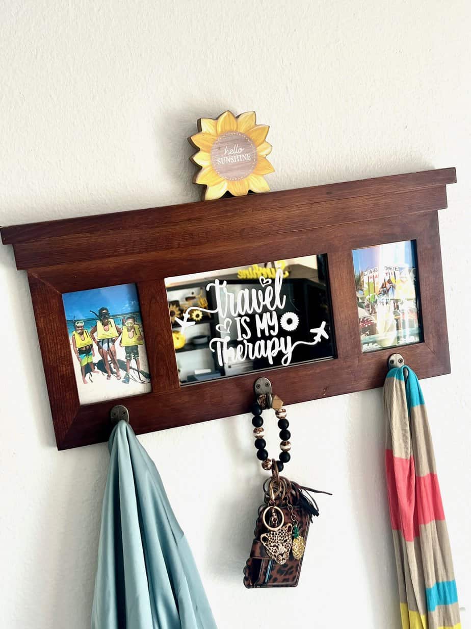 A thrift store coat rack mirror with the words "Travel is my Therapy" etched into it and a family vacation photo on each side of the mirror. The coat rack has a scarf and jacket hanging and a sunflower on top.