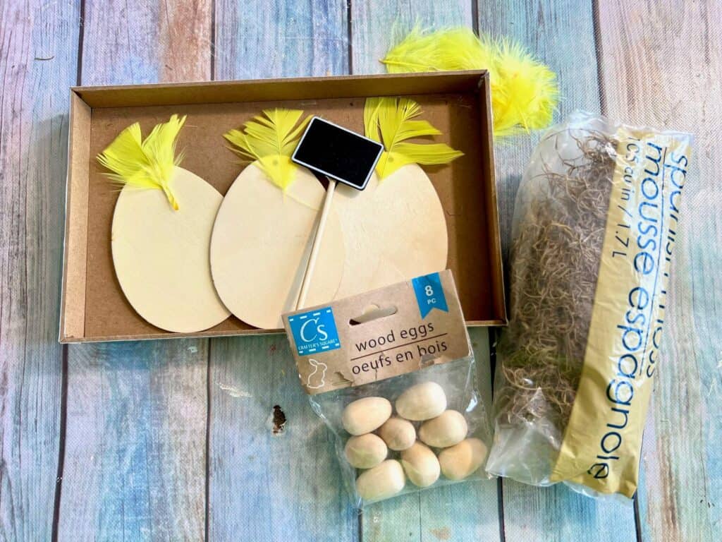 Supplies needed to make the wood easter egg chicks shelf sitter with yellow feathers, wood eggs, spanish moss and other supplies sitting on the table.