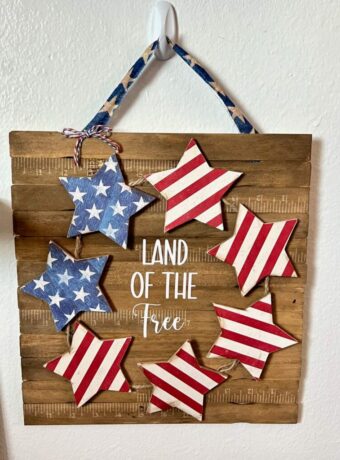 DIY Patriotic Star Wreath Sign with paint sticks background, wood stars in the shape of a wreath with the top left stars blue with white stars and the rest of the stars red and white striped just like the american flag. The center of the sign says "Land of the free". The sign is hanging with a blue star fabric strip.