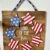 DIY Patriotic Star Wreath Sign with paint sticks background, wood stars in the shape of a wreath with the top left stars blue with white stars and the rest of the stars red and white striped just like the american flag. The center of the sign says "Land of the free". The sign is hanging with a blue star fabric strip.