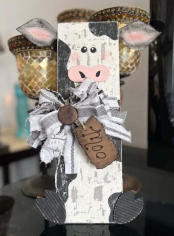 DIY Wood Farmhouse Cow Decor on a chippy cow print rectangular background, fabric ears, big messy black and white fabric bow, and a hangtag that says "Moo".