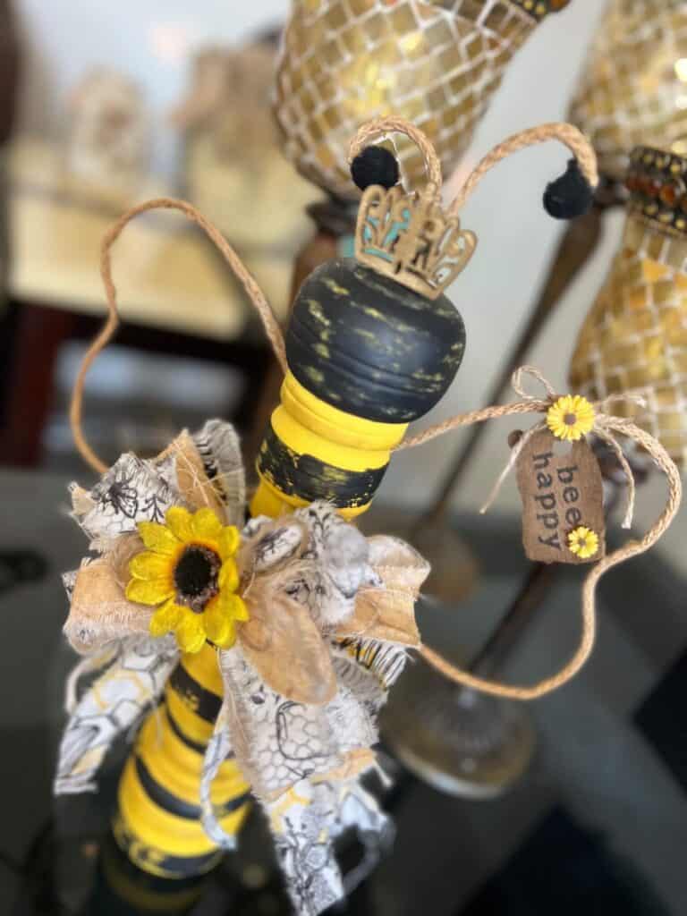Thrift Store Pepper Grinder Queen Bee made from a thrifted wooden pepper grinder yellow with black stripes, wired jute wings with a small tag with sunflowers that says "bee happy", a mini gold crown, and a big messy fabric bow.