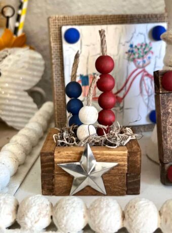 Patriotic Mini Tiered Tray Firecracker Crate with red, white and blue wood bead firecracker with twine and a metal barn star on the front of the crate that is made with Dollar Tree Jenga Blocks and is the perfect size for tiered tray decor.