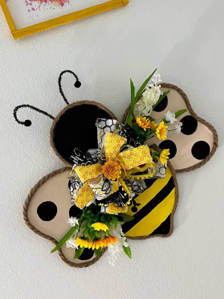 Cardboard Copycat Bumblebee Door Hanger with jute rope around the edge of the cardboard body and a black and yellow stripes with wings with black polka dots and a big messy fabric bow with flowers in the center.