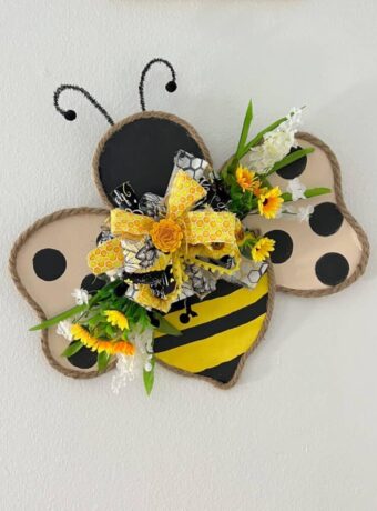 Cardboard Copycat Bumblebee Door Hanger with jute rope around the edge of the cardboard body and a black and yellow stripes with wings with black polka dots and a big messy fabric bow with flowers in the center.