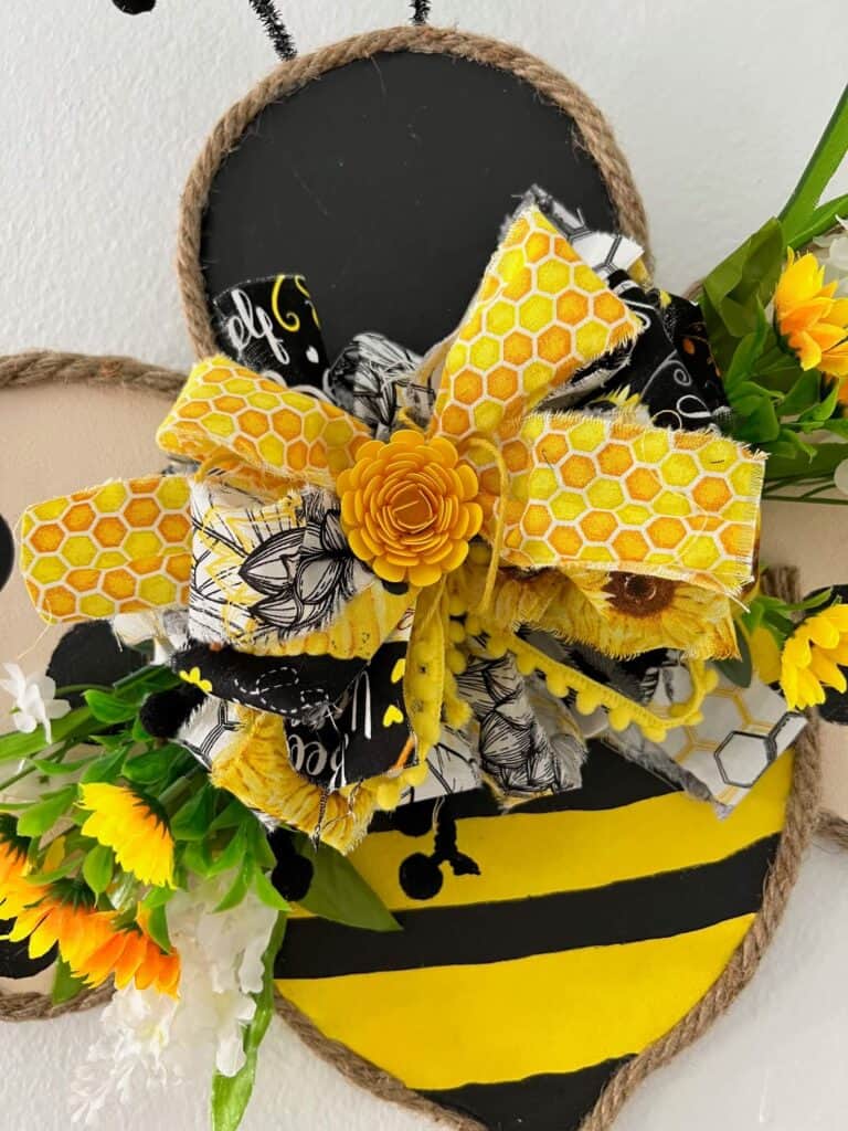 Close up of a big beautiful messy fabric bow with yellow, black, and white fabric, small pom pom ribbon, faux flowers and sunflowers, and a paper flower in the center.