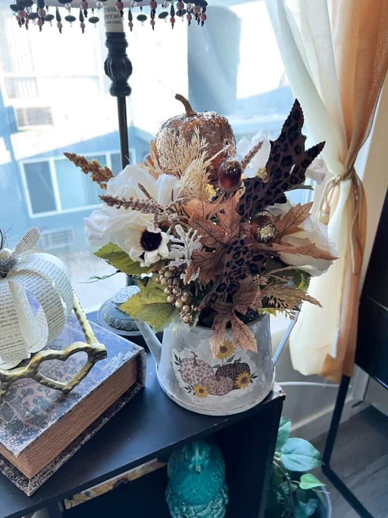 Thrifted watering can flipped into a galvanized metal watering can with a fall floral arrangement, leopard print and gold pumpkin picks, leopard print leaves, and gold acorns, with a leopard print and sunflower pumpkin napkin decoupaged to the front of the watering can for DIY fall centerpiece or home decor.