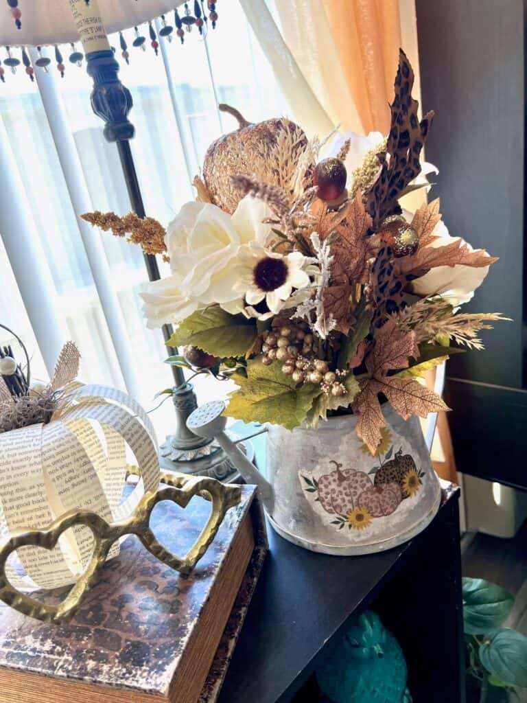 Thrifted Fall Floral Watering can on a bookcase next to a book page pumpkin and gold heat shaped eyeglasses.