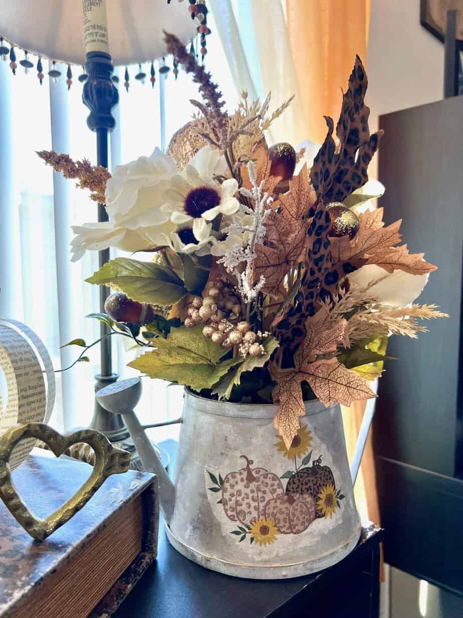 Thrifted watering can flipped into a galvanized metal watering can with a fall floral arrangement, leopard print and gold pumpkin picks, leopard print leaves, and gold acorns, with a leopard print and sunflower pumpkin napkin decoupaged to the front of the watering can for DIY fall centerpiece or home decor.