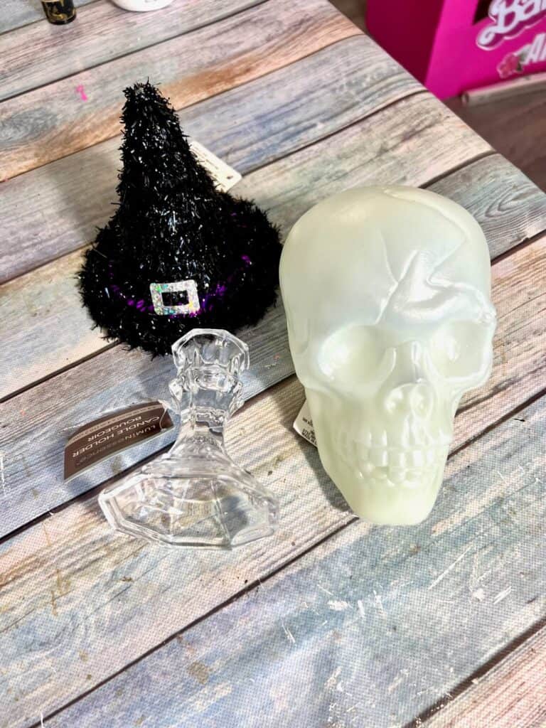 A black tinsel dollar tree witch hat, plastic skull, and glass candlestick sitting on a table.