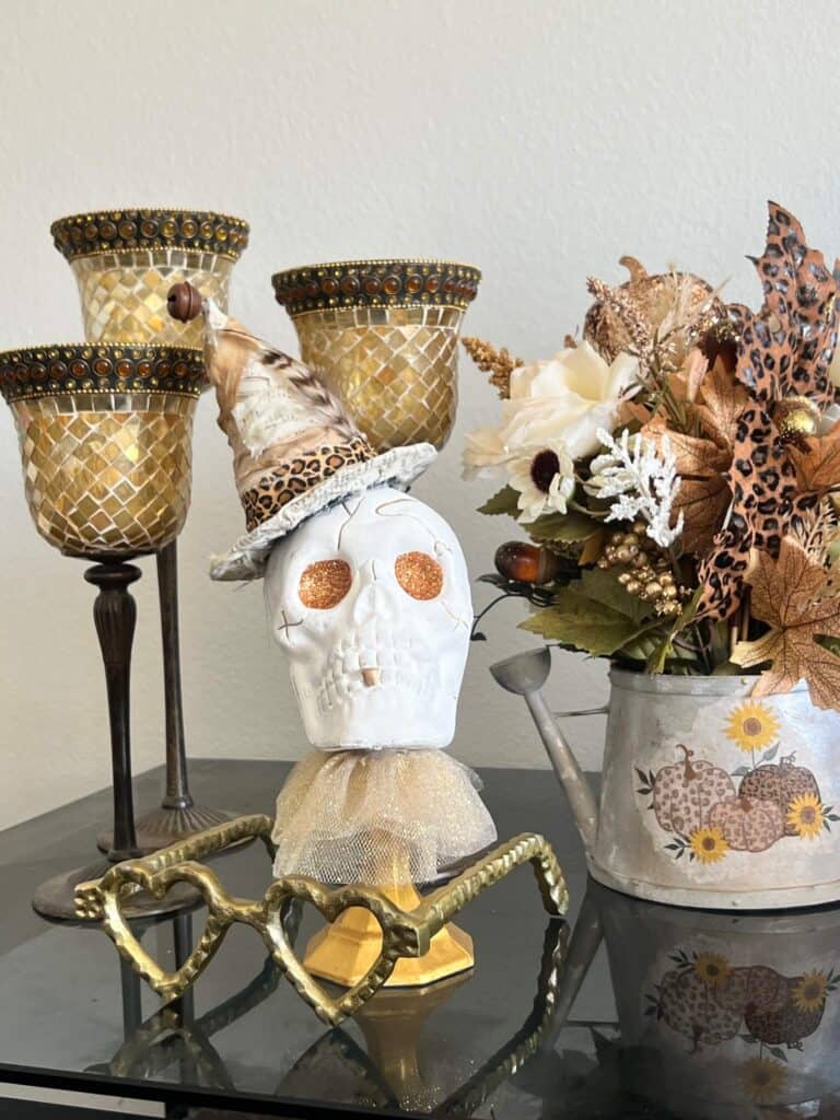 Dollar Tree plastic skull for Halloween, painted white with gold glitter eyes on top of a gold candlestick with a gold tulle neck collar and a witches that was redone with neutral and leopard print fabric and a feather in its hat. With a pair of gold heart shaped eyeglasses sitting nect to it.