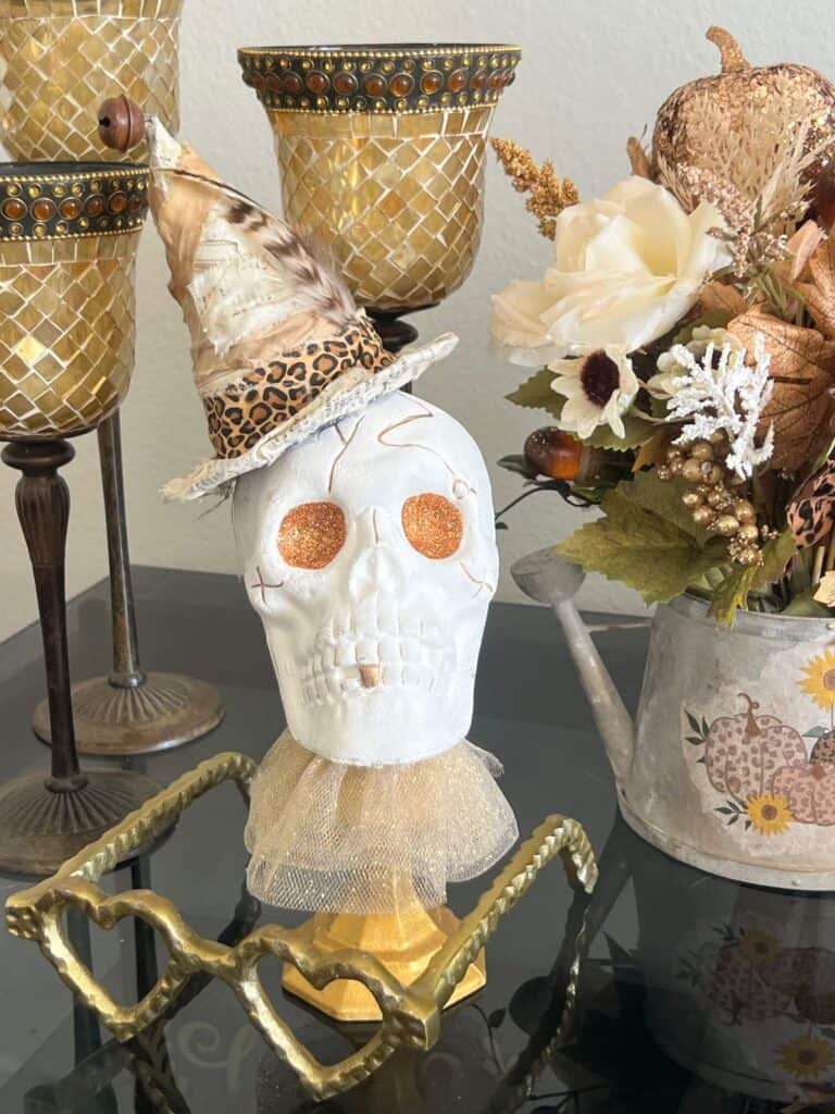 Dollar Tree plastic skull for Halloween, painted white with gold glitter eyes on top of a gold candlestick with a gold tulle neck collar and a witches that was redone with neutral and leopard print fabric and a feather in it's hat.