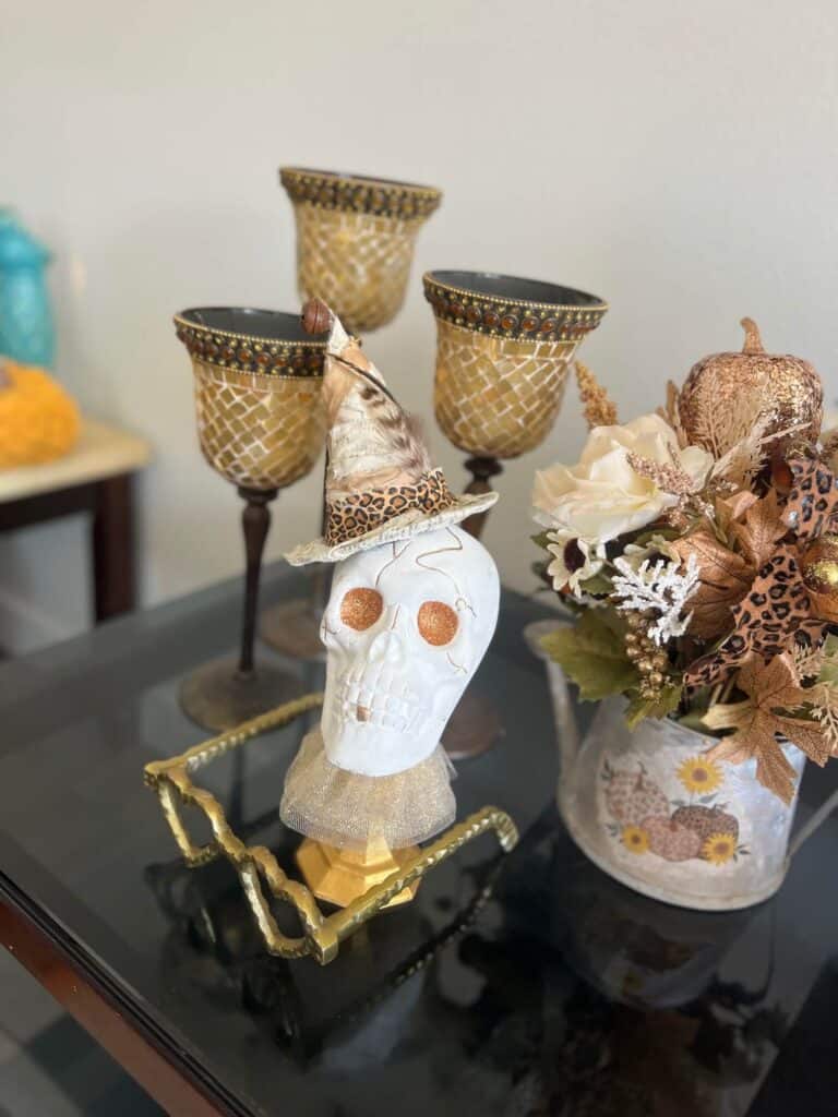 Dollar Tree plastic skull for Halloween, painted white with gold glitter eyes on top of a gold candlestick with a gold tulle neck collar and a witches that was redone with neutral and leopard print fabric and a feather in it's hat.