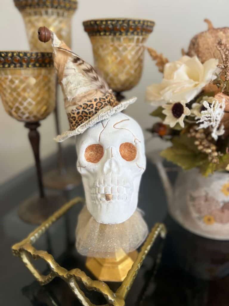 Dollar Tree plastic skull for Halloween, painted white with gold glitter eyes on top of a gold candlestick with a gold tulle neck collar and a witches that was redone with neutral and leopard print fabric and a feather in it's hat.