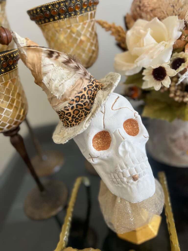 Dollar Tree plastic skull for Halloween, painted white with gold glitter eyes on top of a gold candlestick with a gold tulle neck collar and a witches that was redone with neutral and leopard print fabric and a feather in it's hat.