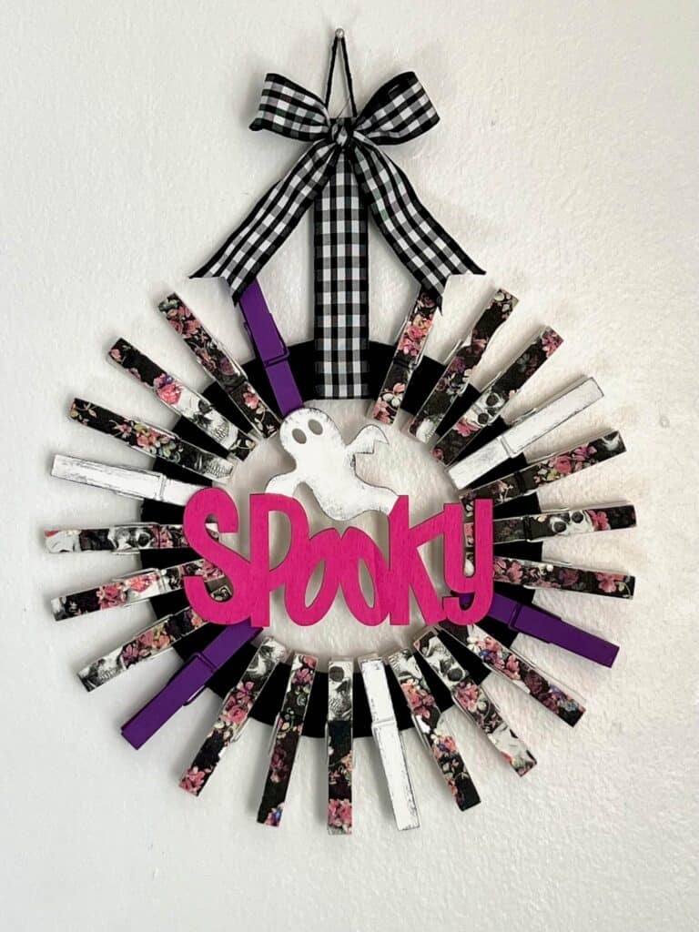 A black cardboard donut shape, with clothespins all around it to make a DIY Halloween wreath shape. The clothes pins are decoupaged pink and purple floral skull napkins, purple and white, and the center is a Dollar Tree Wood cutout of a ghost and the word spooky painted pink, with a buffalo check ribbon hanger.