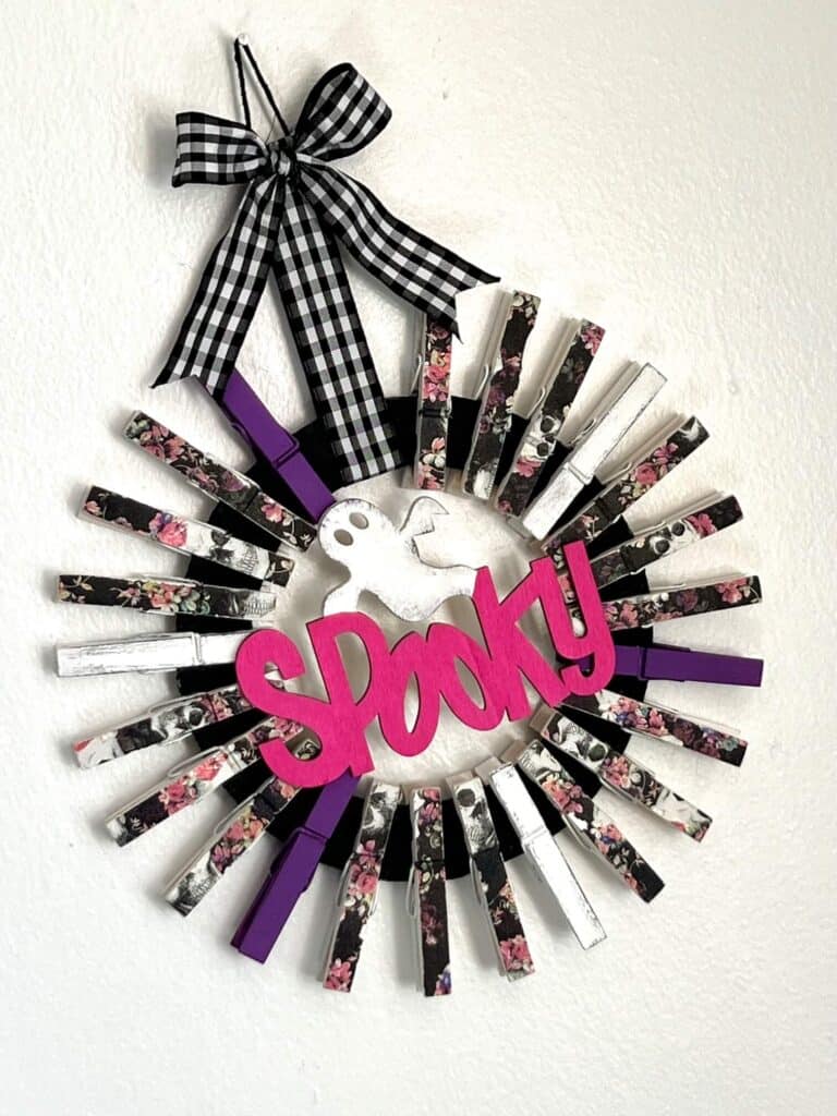 A black cardboard donut shape, with clothespins all around it to make a DIY Halloween wreath shape. The clothes pins are decoupaged pink and purple floral skull napkins, purple and white, and the center is a Dollar Tree Wood cutout of a ghost and the word spooky painted pink, with a buffalo check ribbon hanger.