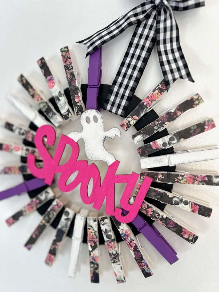 A black cardboard donut shape, with clothespins all around it to make a DIY Halloween wreath shape. The clothes pins are decoupaged pink and purple floral skull napkins, purple and white, and the center is a Dollar Tree Wood cutout of a ghost and the word spooky painted pink, with a buffalo check ribbon hanger.