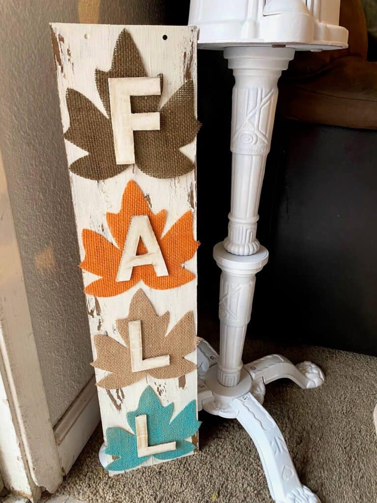 DIY Dollar tree long sign with 4 burlap fall leaves, one brown, orange, tan, and teal, that says FALL. The sign has a white chippy background perfect for decorating your home this autumn season.