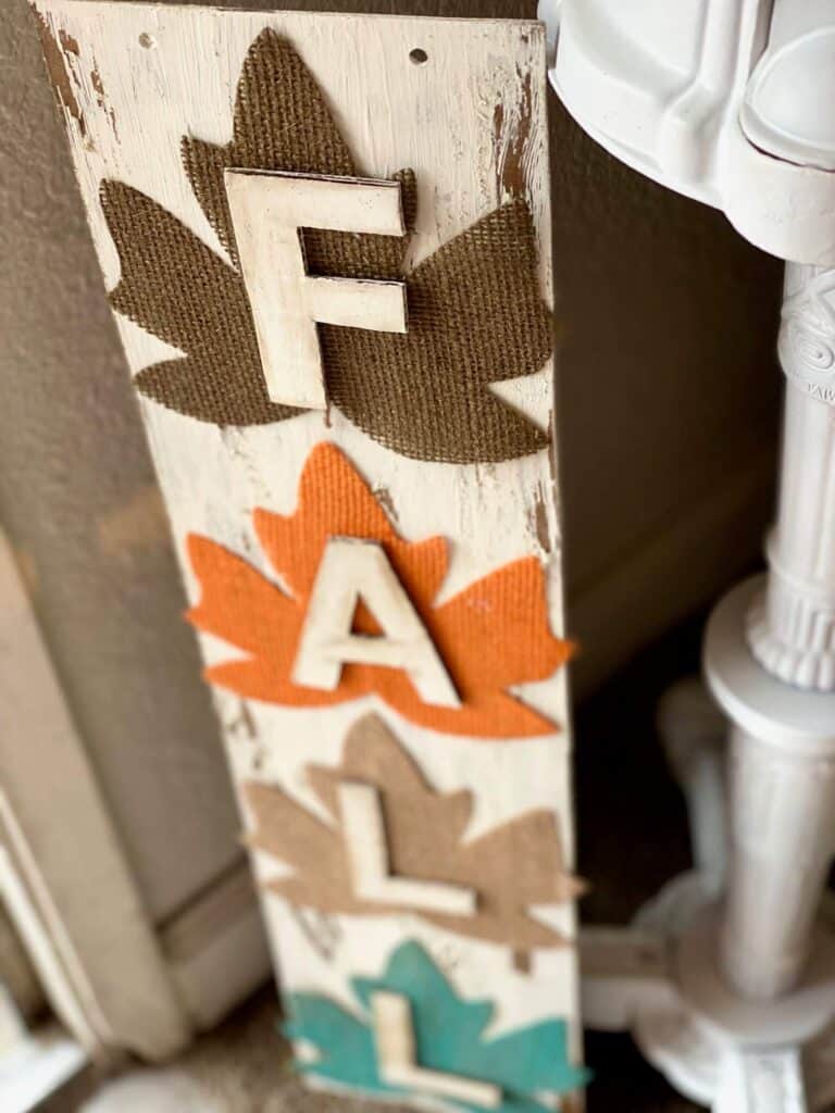 DIY Dollar tree long sign with 4 burlap fall leaves, one brown, orange, tan, and teal, that says FALL. The sign has a white chippy background perfect for decorating your home this autumn season.