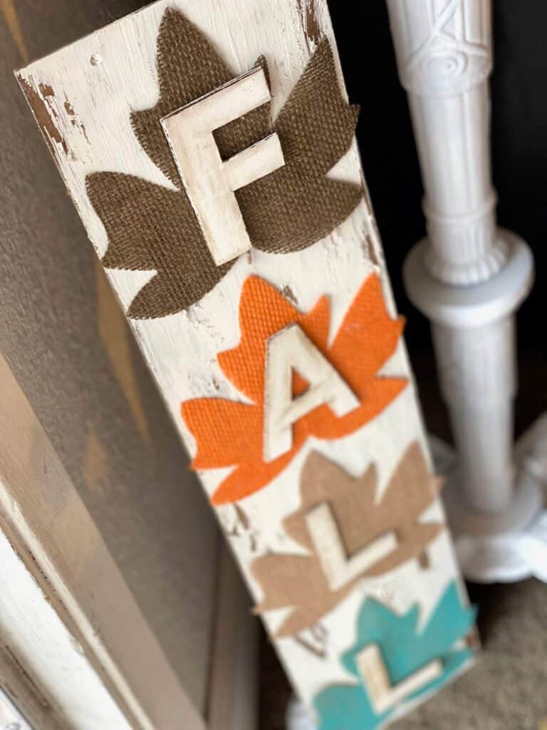 DIY Dollar tree long sign with 4 burlap fall leaves, one brown, orange, tan, and teal, that says FALL. The sign has a white chippy background perfect for decorating your home this autumn season.