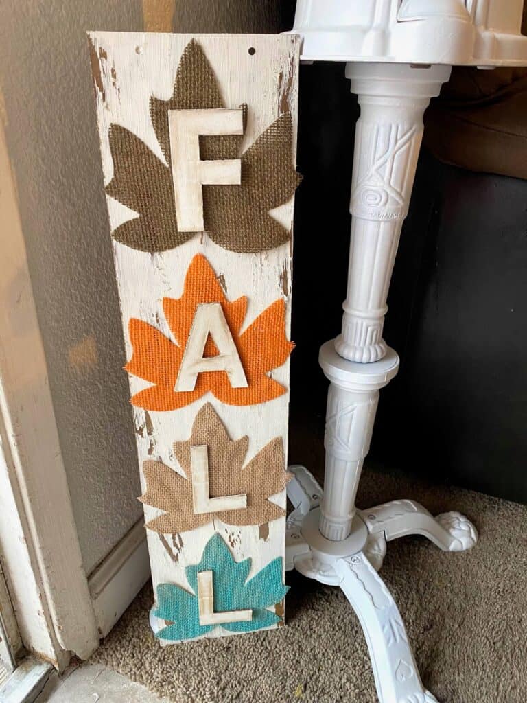 DIY Dollar tree long sign with 4 burlap fall leaves, one brown, orange, tan, and teal, that says FALL. The sign has a white chippy background perfect for decorating your home this autumn season.