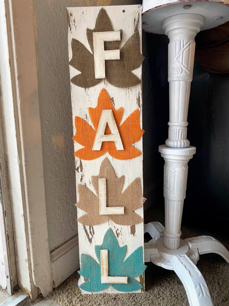 DIY Dollar tree long sign with 4 burlap fall leaves, one brown, orange, tan, and teal, that says FALL. The sign has a white chippy background perfect for decorating your home this autumn season.