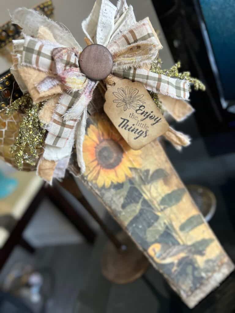 DIY Scrap wood sunflower decor with a free sunflower printable on rice paper, decoupaged onto a piece of scrap wood and a big messy fabric bow with a hangtag that says "enjoy the little things".