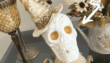 Dollar Tree plastic skull for Halloween, painted white with gold glitter eyes on top of a gold candlestick with a gold tulle neck collar and a witches that was redone with neutral and leopard print fabric and a feather in it