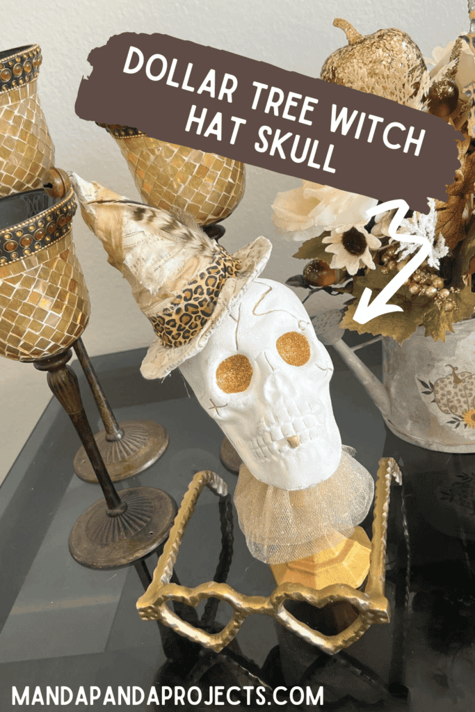 Dollar Tree plastic skull for Halloween, painted white with gold glitter eyes on top of a gold candlestick with a gold tulle neck collar and a witches that was redone with neutral and leopard print fabric and a feather in it's hat.