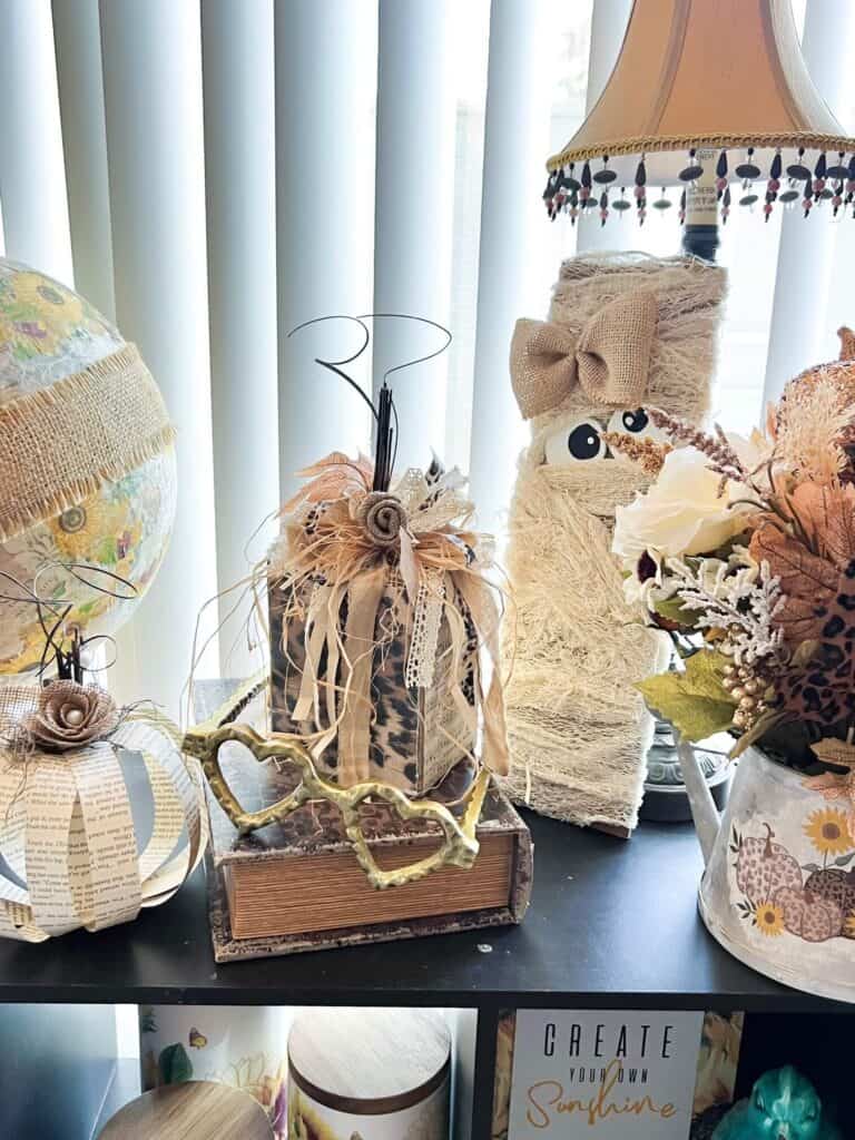 Neutral wood pumpkin craft on a bookshelf with a mummy, a book page pumpkin, and a pair of gold heart glasses.