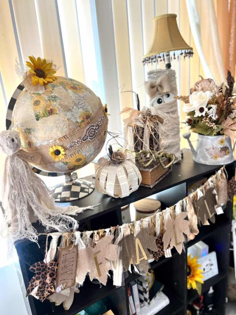 Neutral wood pumpkin craft on a bookshelf with a mummy, a book page pumpkin, a rag ghost, a sunflower world globe, a watering can floral arrangement, and a pair of gold heart glasses.