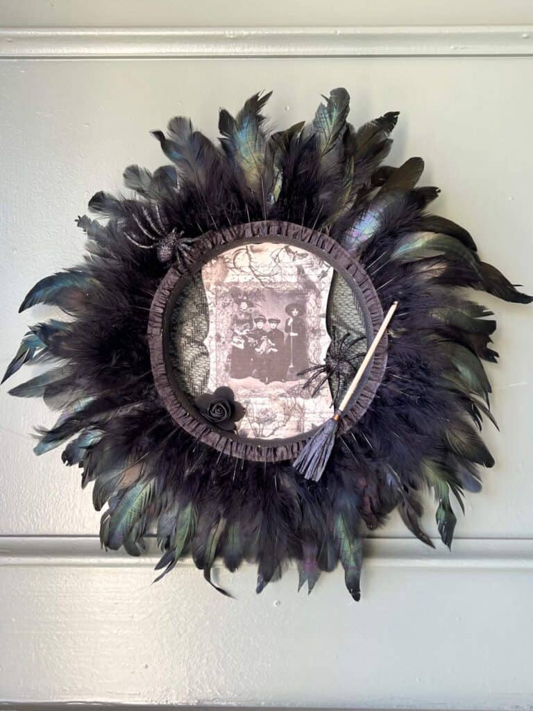 Dollar Tree styrofoam wreath form with black feather trim around the outside, vintage witches rice paper sign in the center, and embellished with faux spiders, mini broomstick, and a black rose for DIY Halloween crafts and decor.