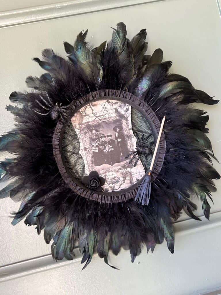 Dollar Tree styrofoam wreath form with black feather trim around the outside, vintage witches rice paper sign in the center, and embellished with faux spiders, mini broomstick, and a black rose for DIY Halloween crafts and decor.