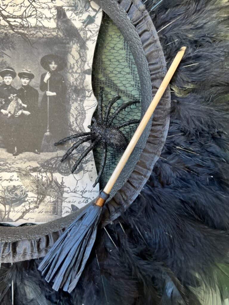 A black sparkly spider and a mini broomstick glued to the bottom right of the Halloween Feather Wreath.