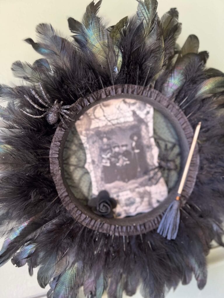 Dollar Tree styrofoam wreath form with black feather trim around the outside, vintage witches rice paper sign in the center, and embellished with faux spiders, mini broomstick, and a black rose for DIY Halloween crafts and decor.