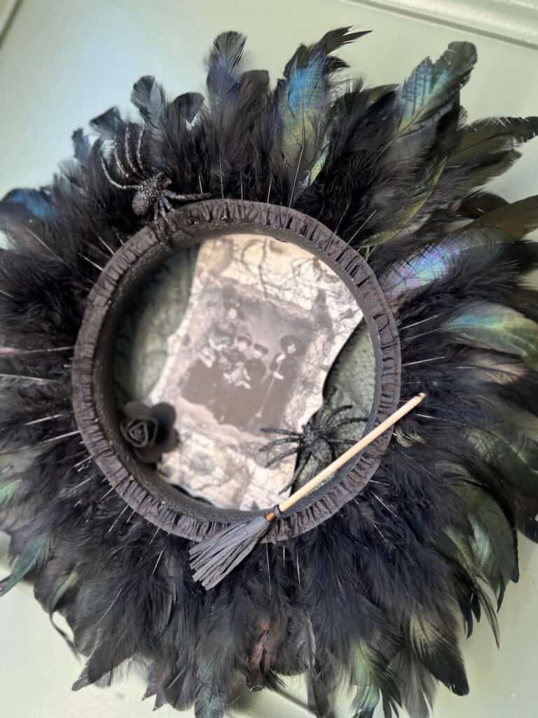 Dollar Tree styrofoam wreath form with black feather trim around the outside, vintage witches rice paper sign in the center, and embellished with faux spiders, mini broomstick, and a black rose for DIY Halloween crafts and decor.