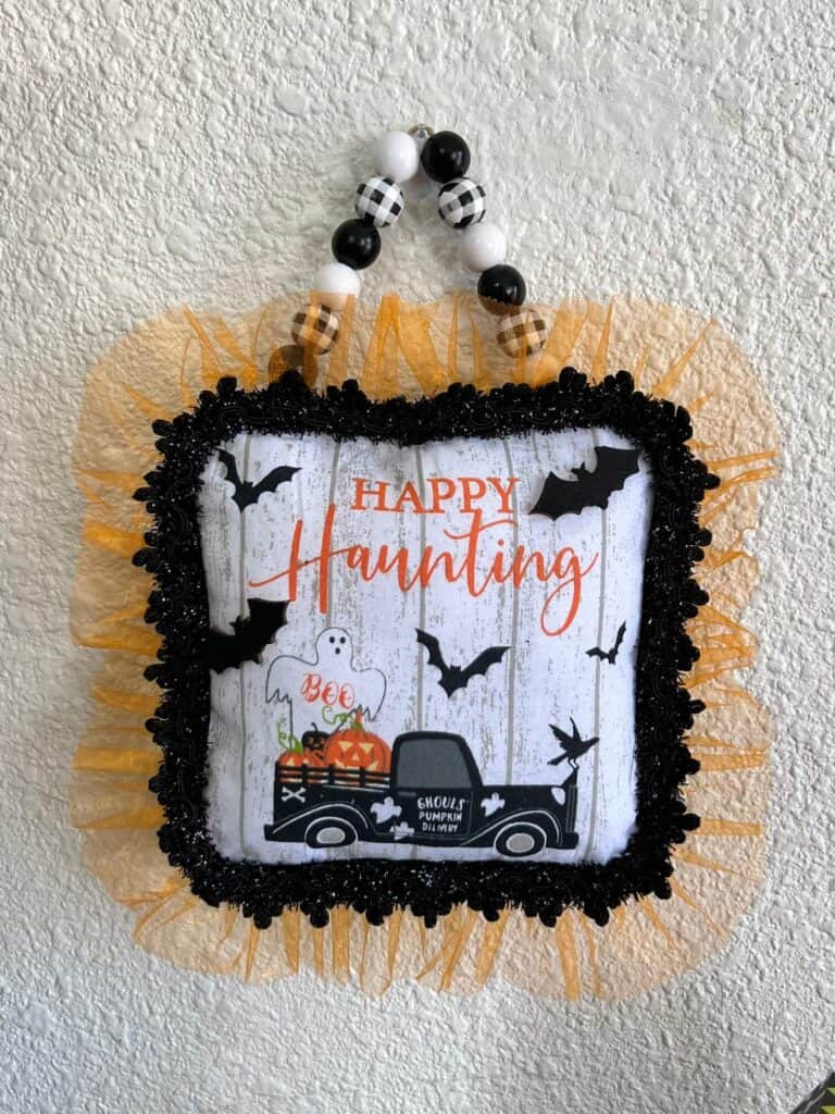 Dollar Tree Halloween Pot Holder with a Ghost, bats, and an old truck that says "Happy Haunting with orange tulle trim around the edge and a black and white buffalo check wood bead hanger, DIY doorknob hanger.