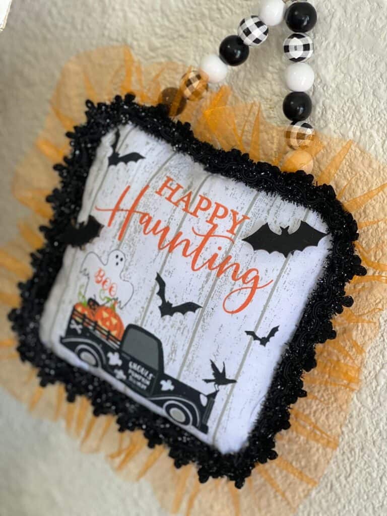 Dollar Tree Halloween Pot Holder with a Ghost, bats, and an old truck that says "Happy Haunting with orange tulle trim around the edge and a black and white buffalo check wood bead hanger, DIY doorknob hanger.