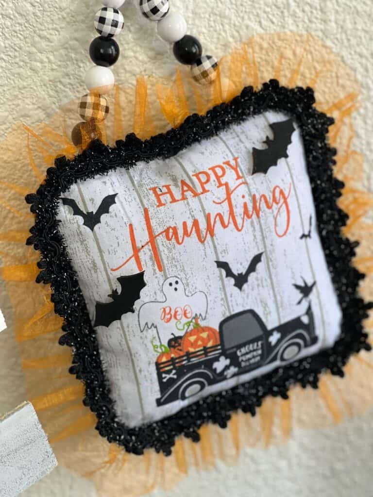 Dollar Tree Halloween Pot Holder with a Ghost, bats, and an old truck that says "Happy Haunting with orange tulle trim around the edge and a black and white buffalo check wood bead hanger, DIY doorknob hanger.