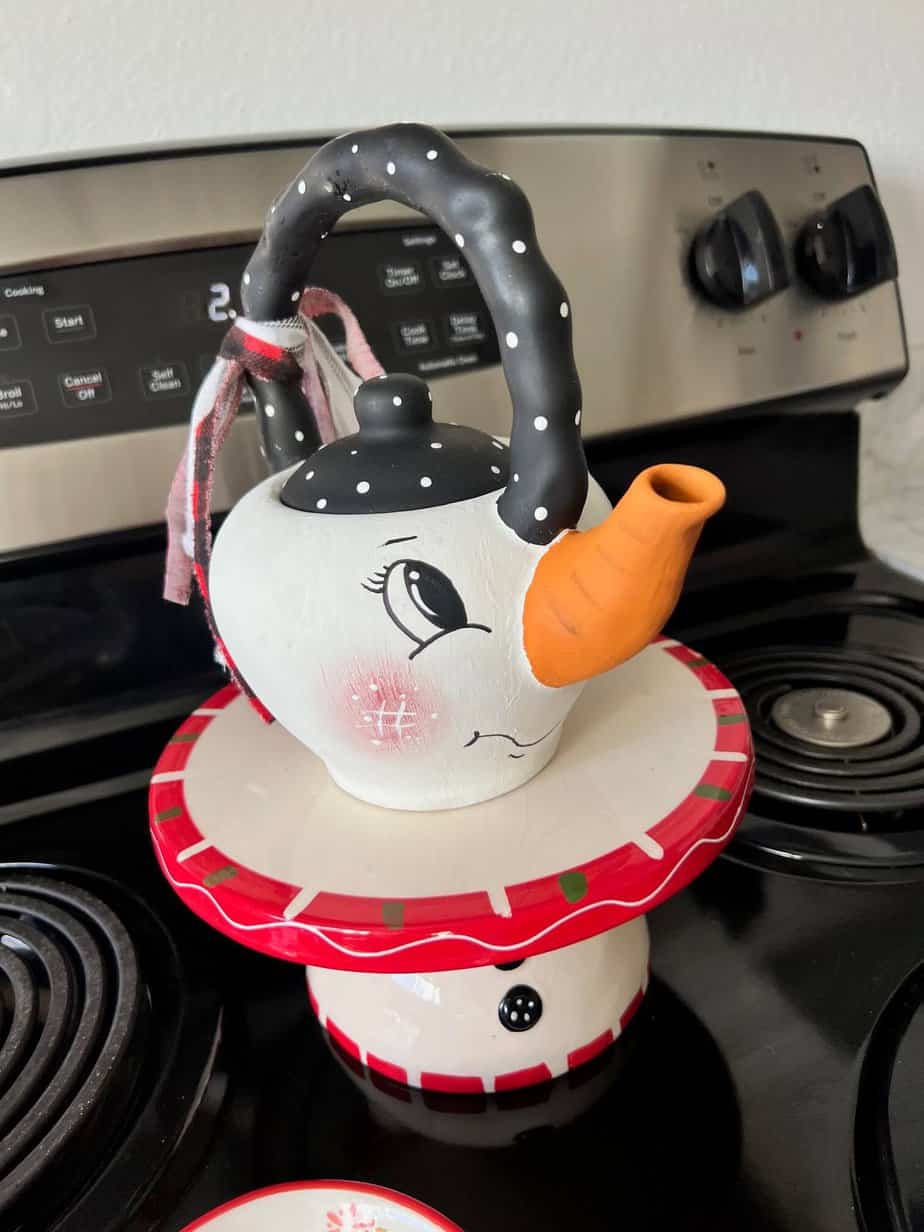 Thrifted Hand painted snowman teapot with the "spout" painted orange as the "carrot" nose, big black eyed, rosy pink cheeks, black and white polka dot handle, and a black, red, and white scrappy fabric bow on the handle.