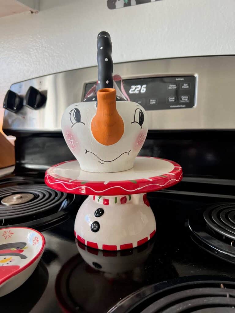 Thrifted Hand painted snowman teapot with the "spout" painted orange as the "carrot" nose, big black eyed, rosy pink cheeks, black and white polka dot handle, and a black, red, and white scrappy fabric bow on the handle.