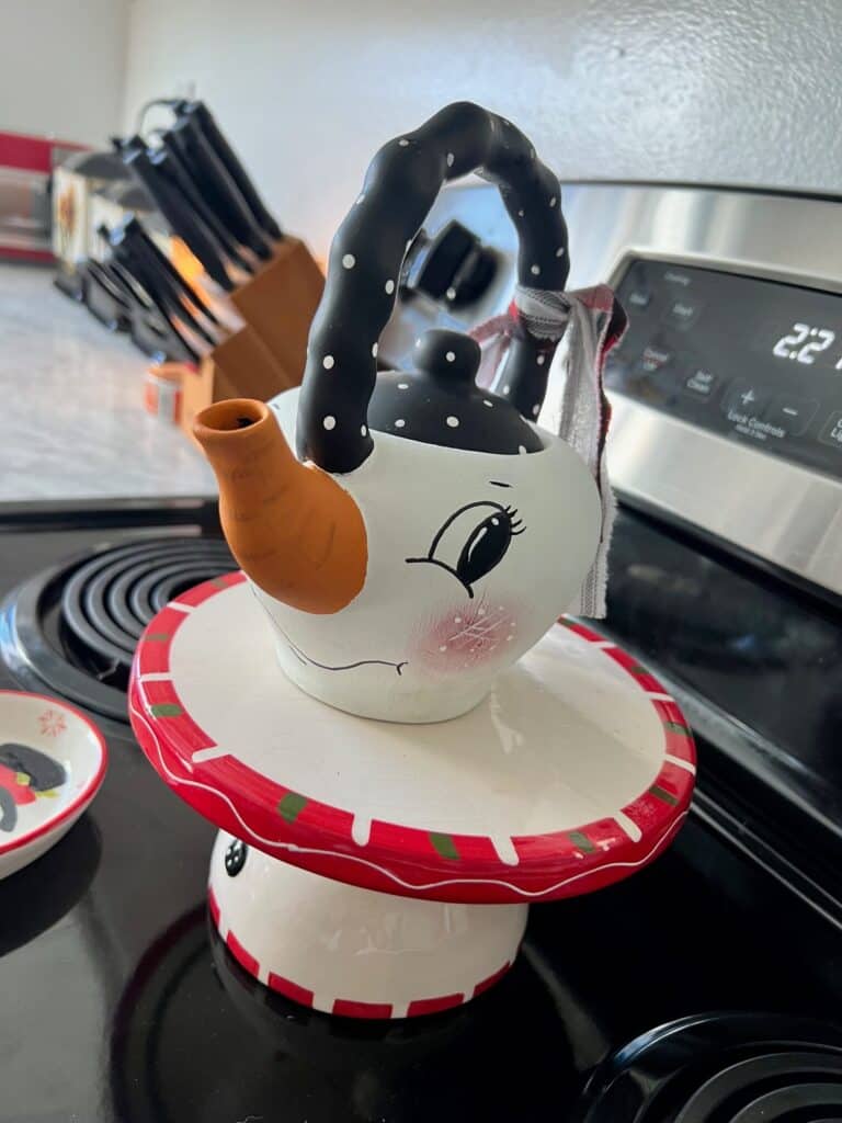 Thrifted Hand painted snowman teapot with the "spout" painted orange as the "carrot" nose, big black eyed, rosy pink cheeks, black and white polka dot handle, and a black, red, and white scrappy fabric bow on the handle.