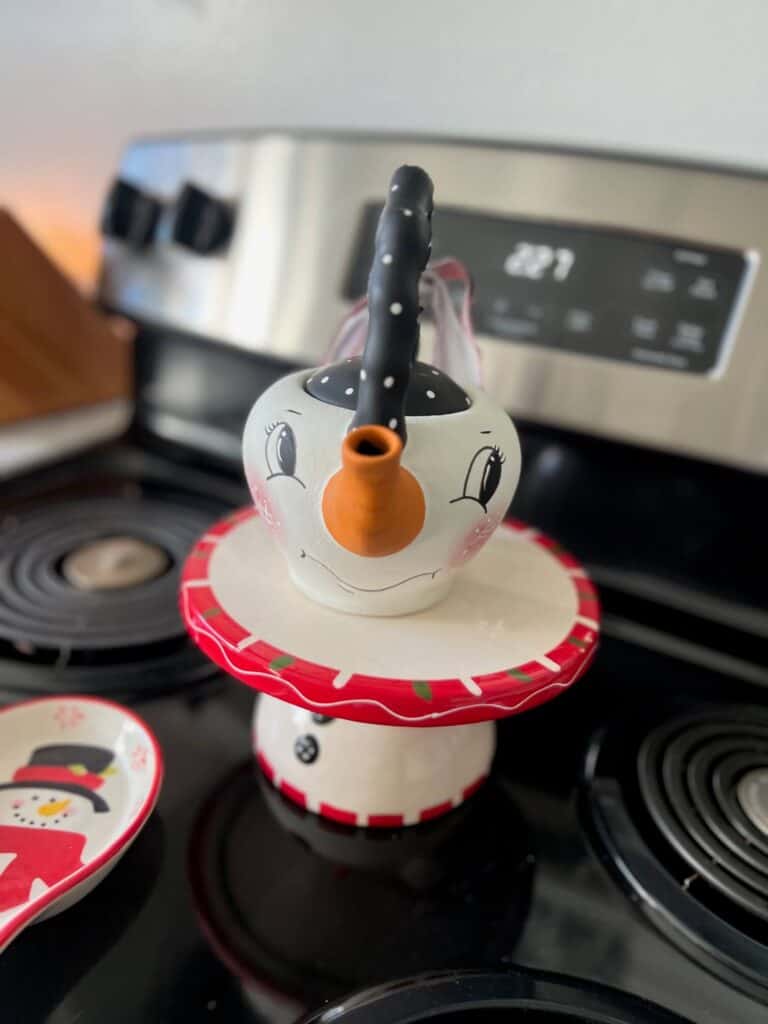 Thrifted Hand painted snowman teapot with the "spout" painted orange as the "carrot" nose, big black eyed, rosy pink cheeks, black and white polka dot handle, and a black, red, and white scrappy fabric bow on the handle.